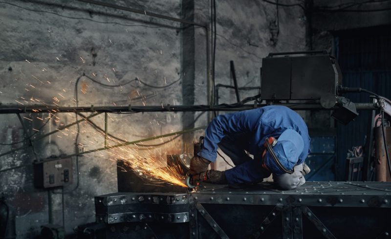 Front view of fitter in special blue uniform working with electric steel cutter machine for welding special metal. Concept of process working with grinding machine in garage.